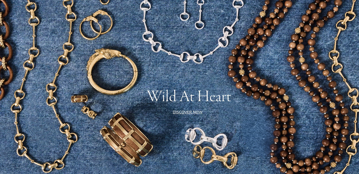 A styled flat-lay of various jewelry pieces, including gold and silver heart-link chains, rings, earrings, and beaded necklaces, arranged on a textured blue denim background. The text 'Wild At Heart' and 'DISCOVER NOW' are printed in the center.