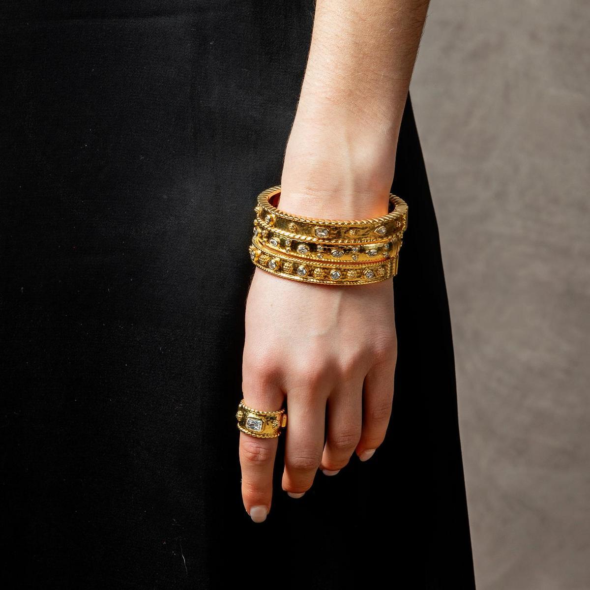 A close-up of a person's hand and wrist wearing a stack of ornate gold bangles and a matching gold ring with green stone accents, set against a black dress and a neutral gray background.