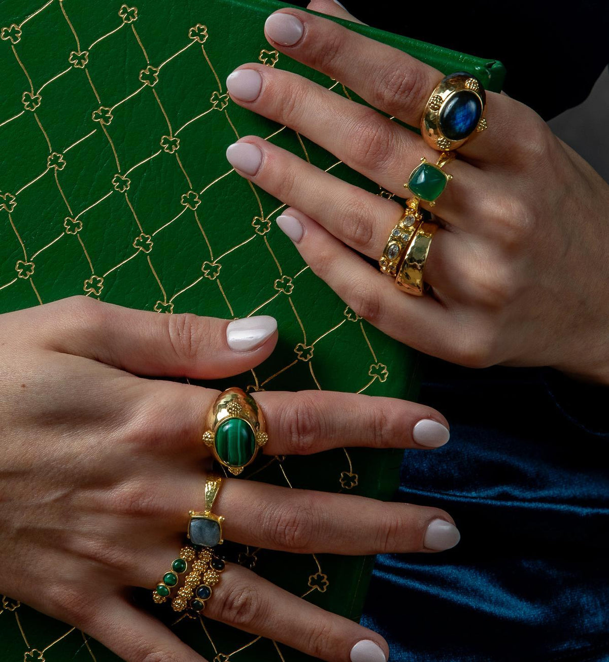 Close-up of two hands wearing multiple gold rings with green and blue stones, posed against a green patterned box and dark blue fabric.
