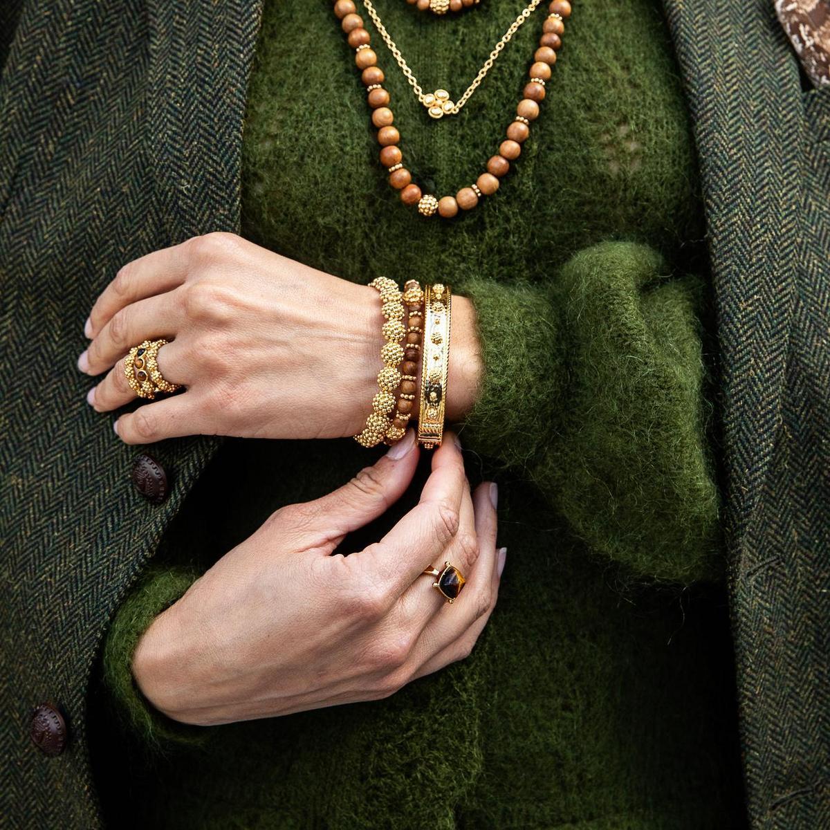 Close-up of a person wearing layered gold jewelry, including a bangle bracelet, rings, and beaded necklaces, styled with an olive green knit sweater and a dark herringbone coat.