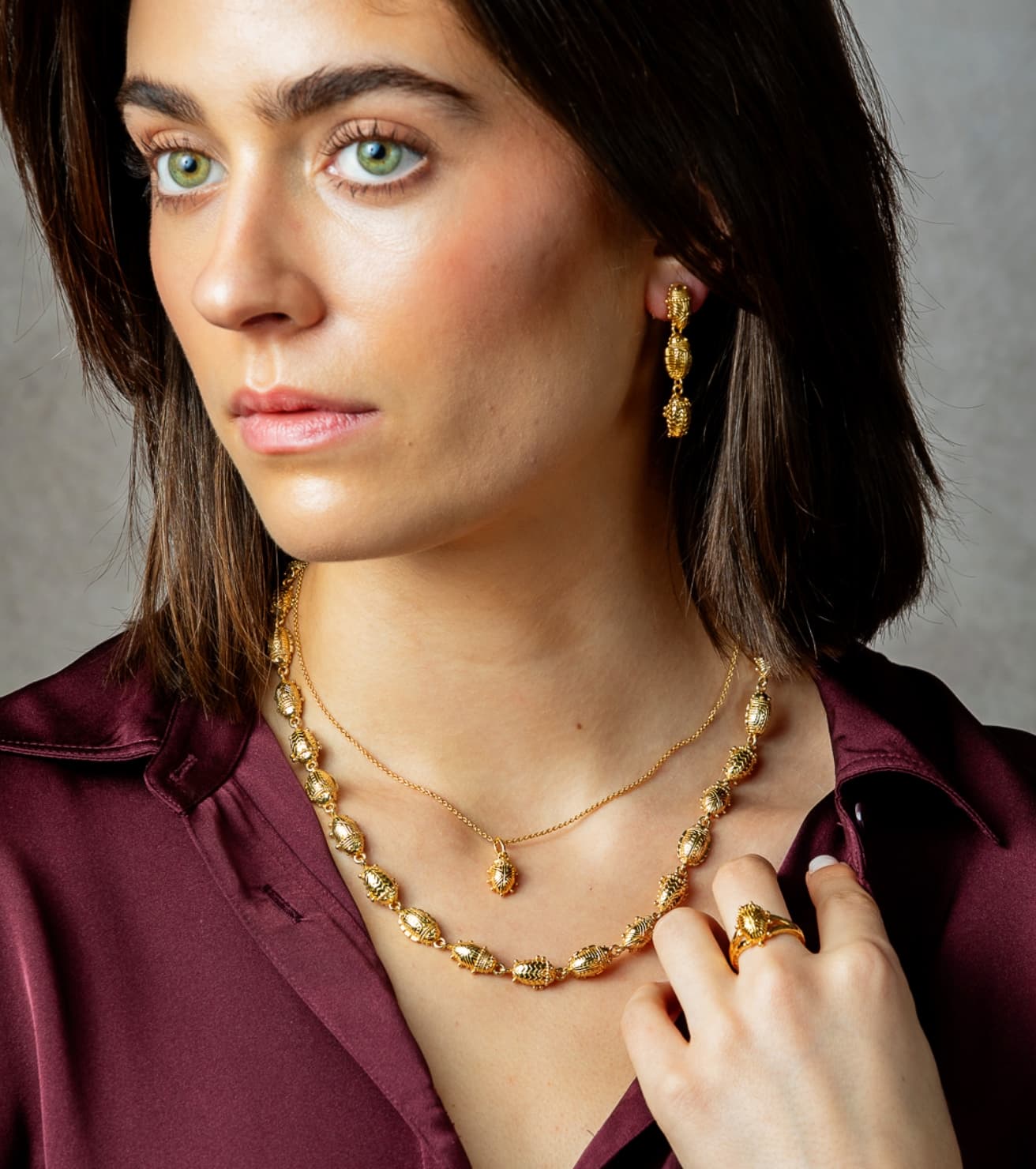 A close-up of a woman wearing gold jewelry, including a beaded necklace, a delicate chain necklace with a small pendant, and a matching drop earring, paired with a dark burgundy satin shirt against a neutral gray background.