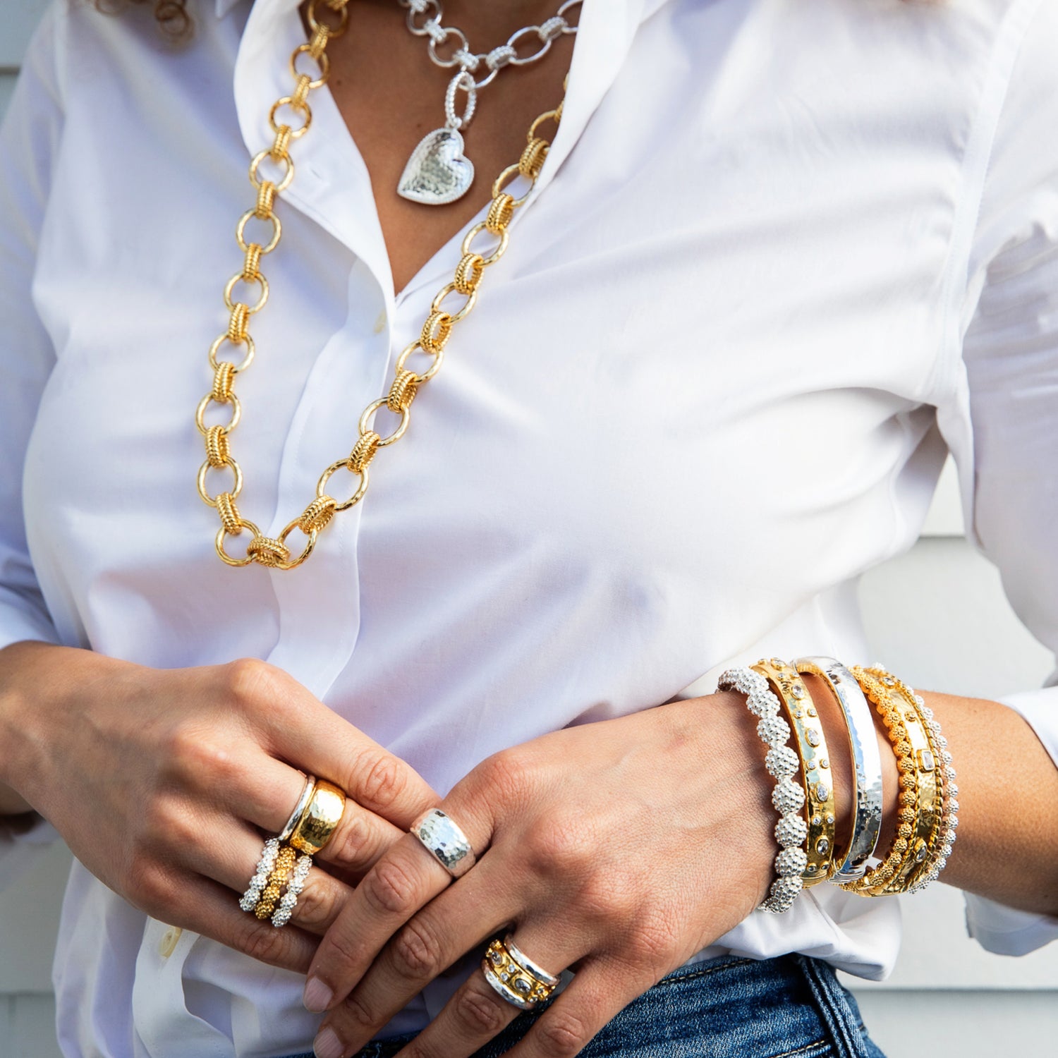 A close-up of a person wearing layered jewelry, including a chunky gold chain necklace, a silver heart pendant necklace, multiple gold and silver bracelets on one wrist, and several rings on both hands, styled with a white button-down shirt and blue jeans.