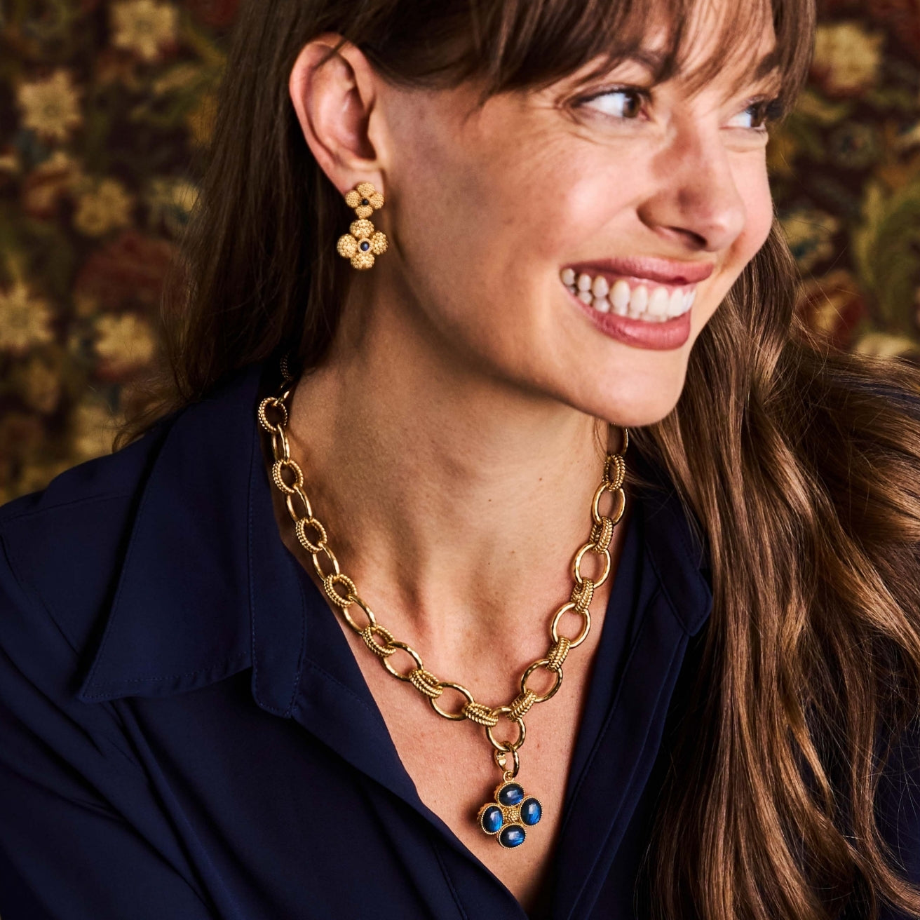 A close-up lifestyle shot of a woman wearing a gold chain necklace with a blue and gold berry pendant, matching gold flower earrings, and a navy blue shirt against a warm, patterned background.