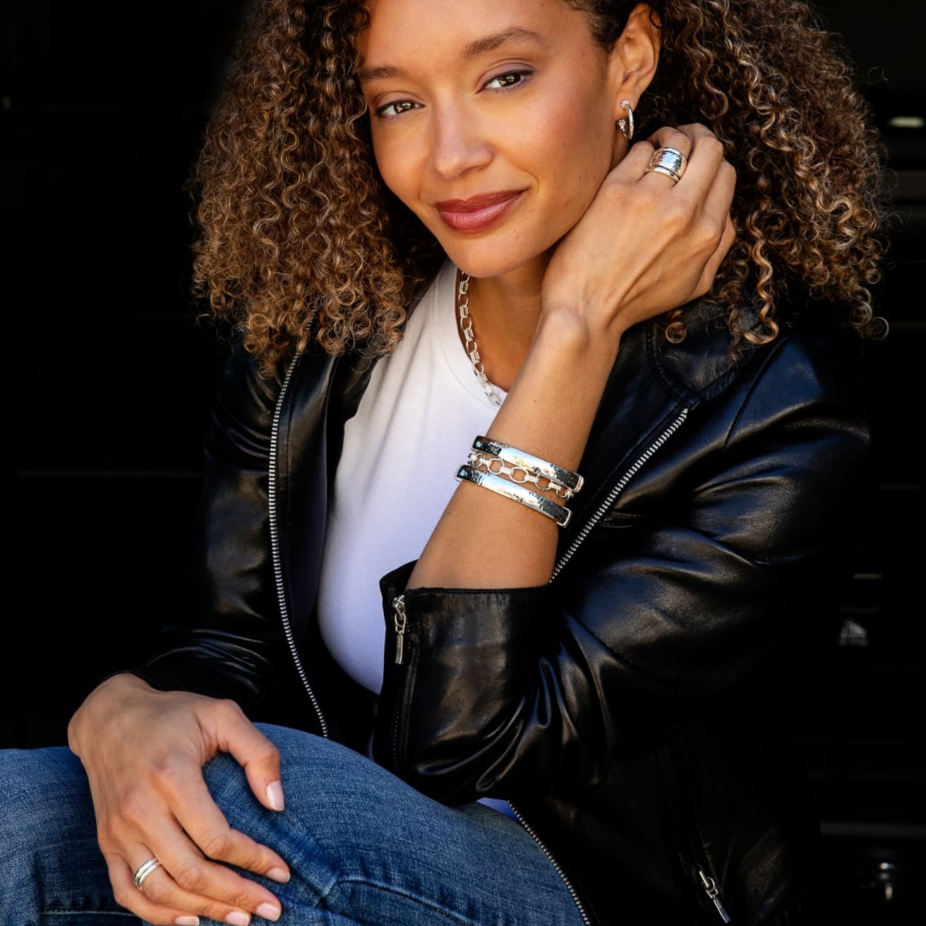 A woman with curly hair wearing a black leather jacket, white t-shirt, and blue jeans, accessorized with silver rings and bracelets, posing against a dark background.