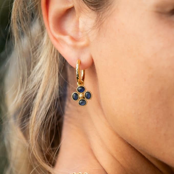 A close-up of a woman's ear wearing a gold huggie hoop earring with a dangling floral cluster of dark blue round stones.