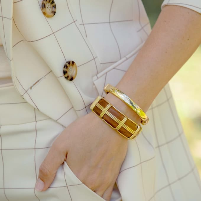 A close-up of a person's wrist wearing two gold-toned bangle bracelets, one with a brown geometric grid pattern and the other with a textured yellow-green finish, styled with a cream-colored checkered blazer.