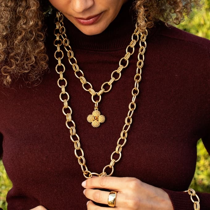 A close-up of a model wearing a double-layered gold chain necklace with a textured flower-shaped pendant, styled with a dark burgundy turtleneck sweater.