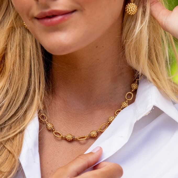 A close-up lifestyle shot of a woman wearing a textured gold chain necklace and matching gold dangle earrings, styled with a white collared shirt.