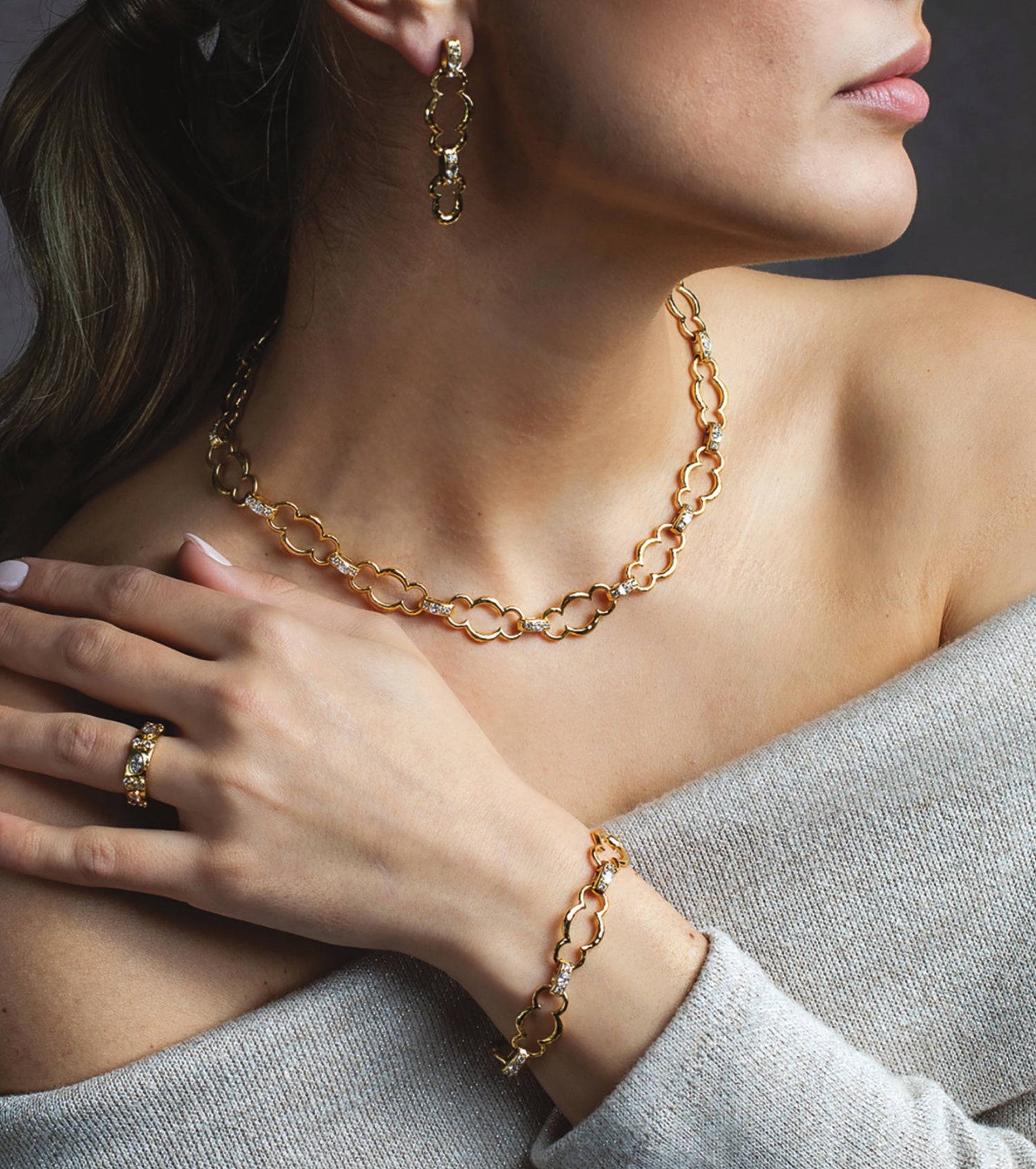 A close-up lifestyle shot of a model wearing a gold jewelry set, including a link chain necklace, matching drop earrings, a ring, and a bracelet, styled with a gray off-the-shoulder sweater.