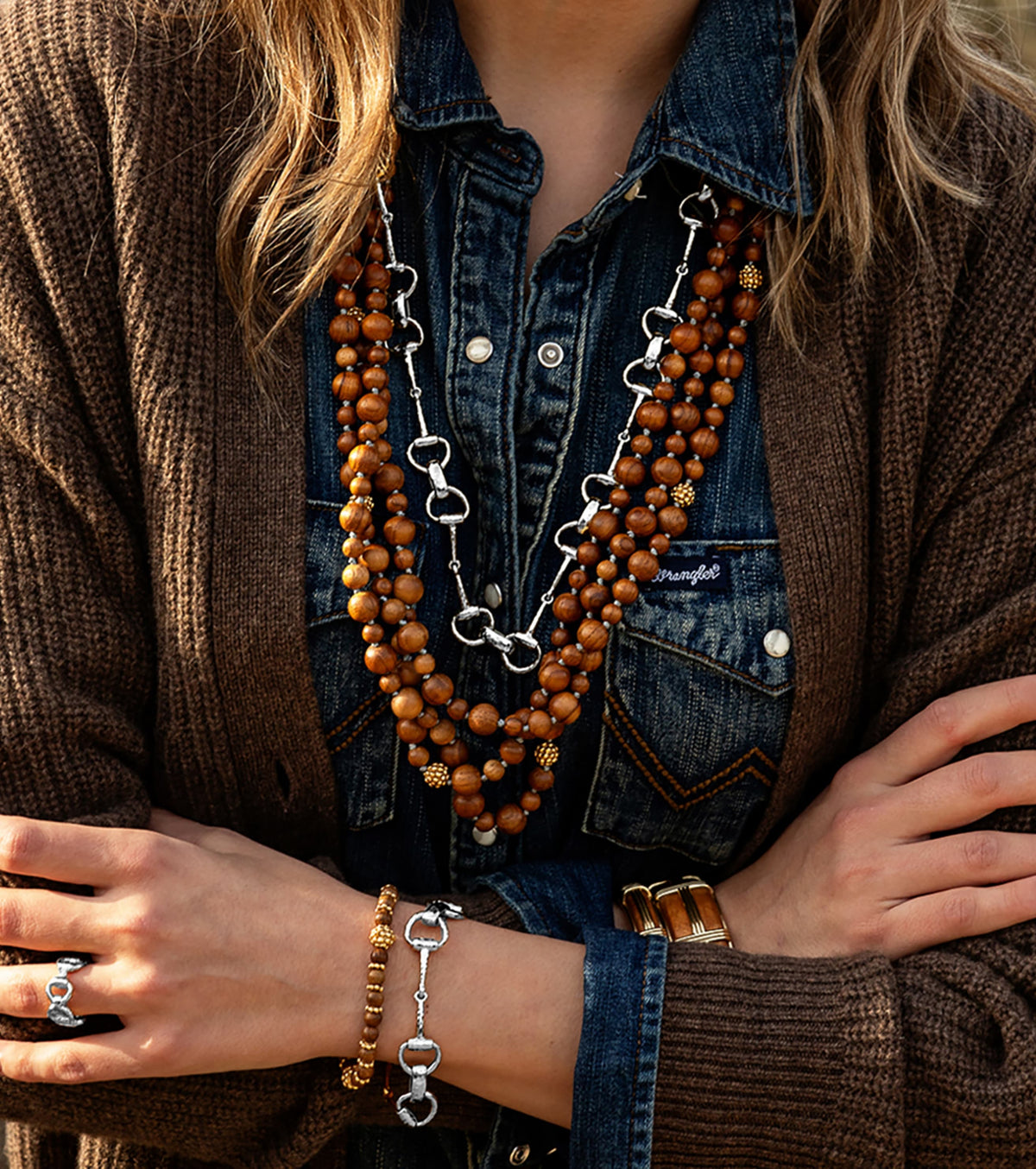 Lifestyle image of women wearing denim shirt with brown sweater, silver equestrian snaffle bit jewelry and teak beaded jewelry necklace and bracelet