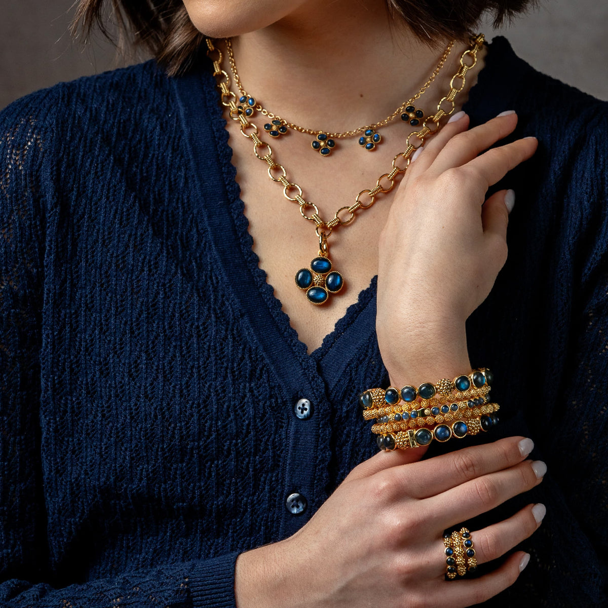 A close-up of a person wearing layered gold-tone jewelry, including a necklace with blue and gold floral motifs, a matching bracelet stack, and a ring, styled with a dark navy cardigan.