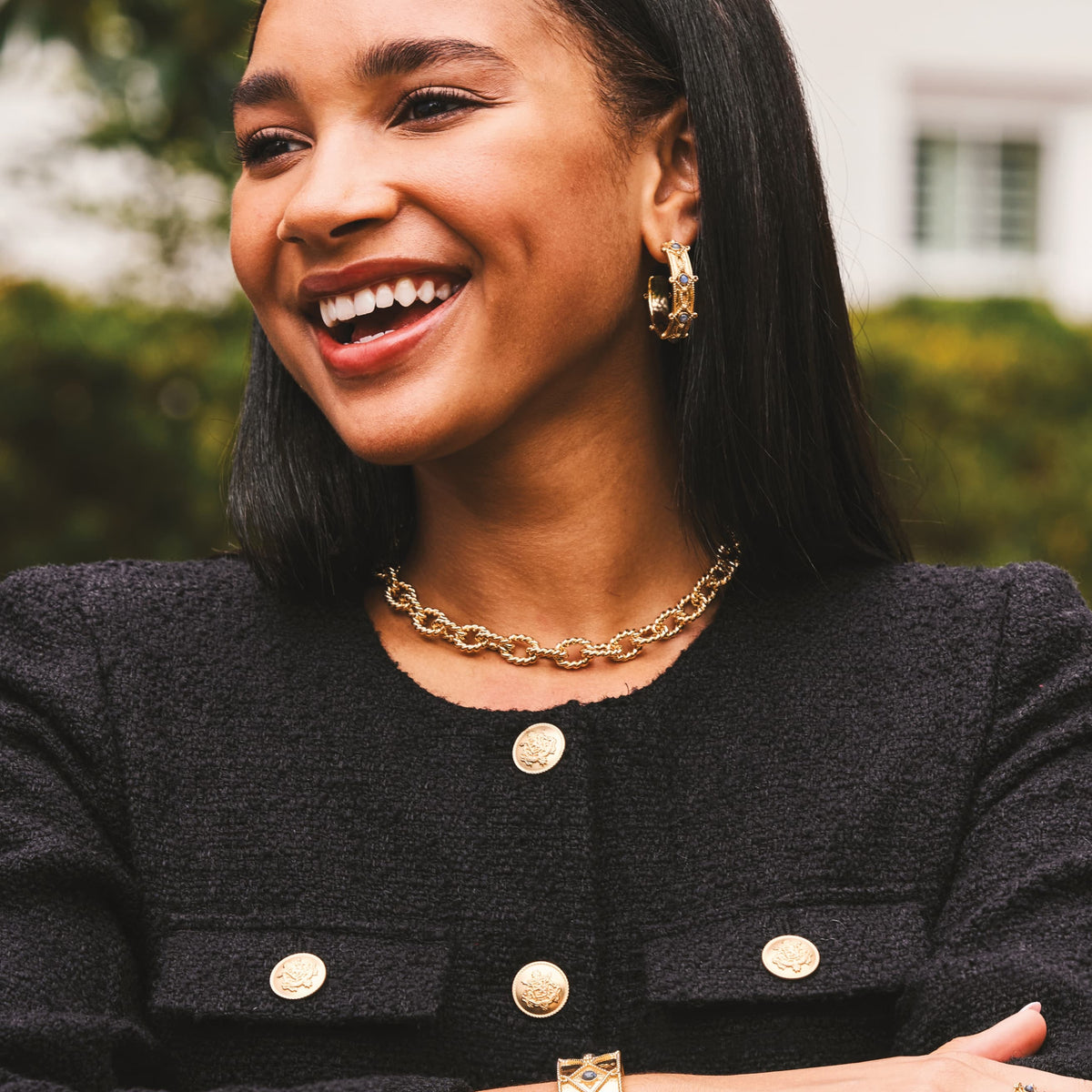 A smiling woman with dark hair wearing a black textured jacket with gold buttons, a gold chain necklace, and gold hoop earrings, photographed outdoors with a blurred background.