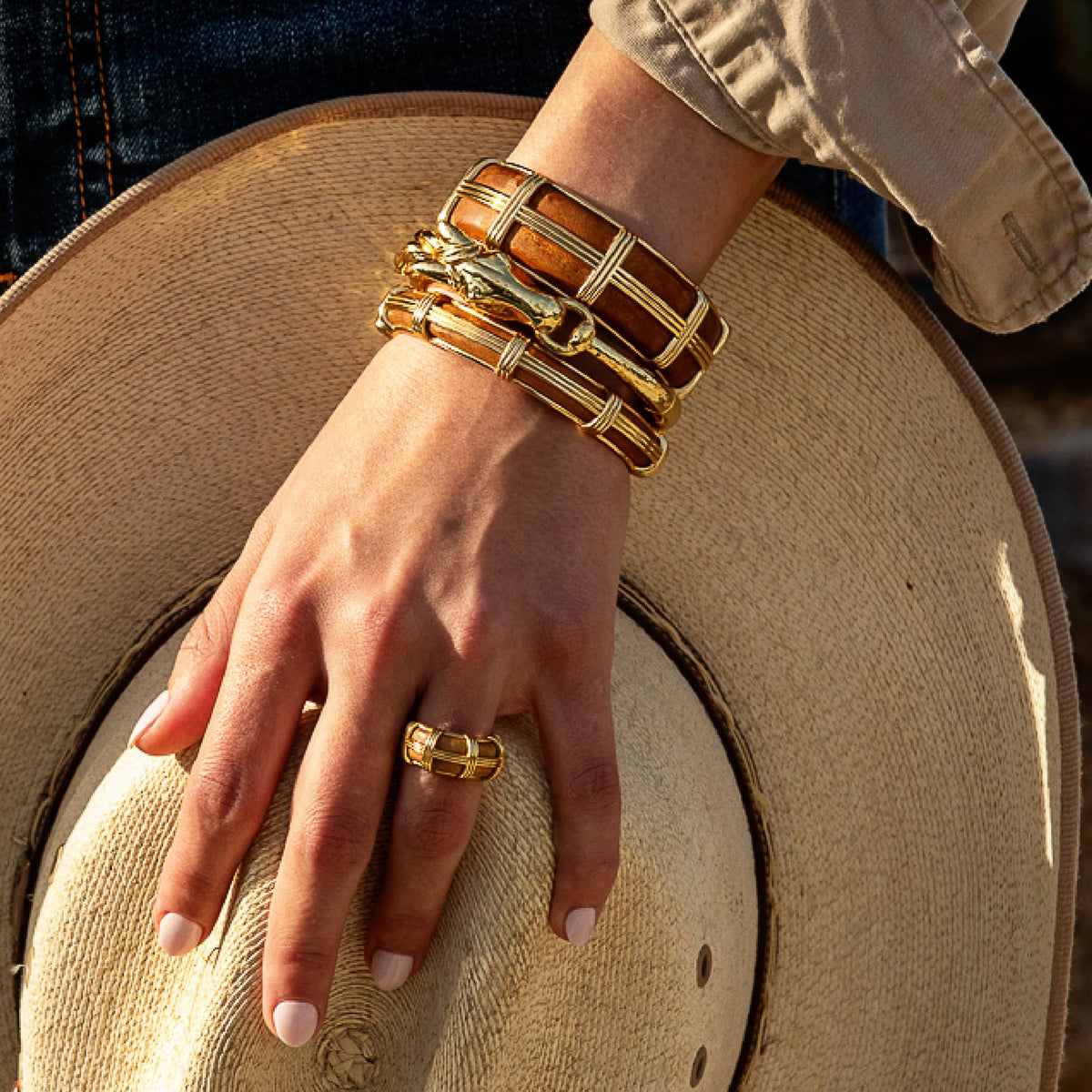 A close-up of a person's hand wearing stacked gold and brown bangles and a matching gold ring, resting on a beige straw hat.
