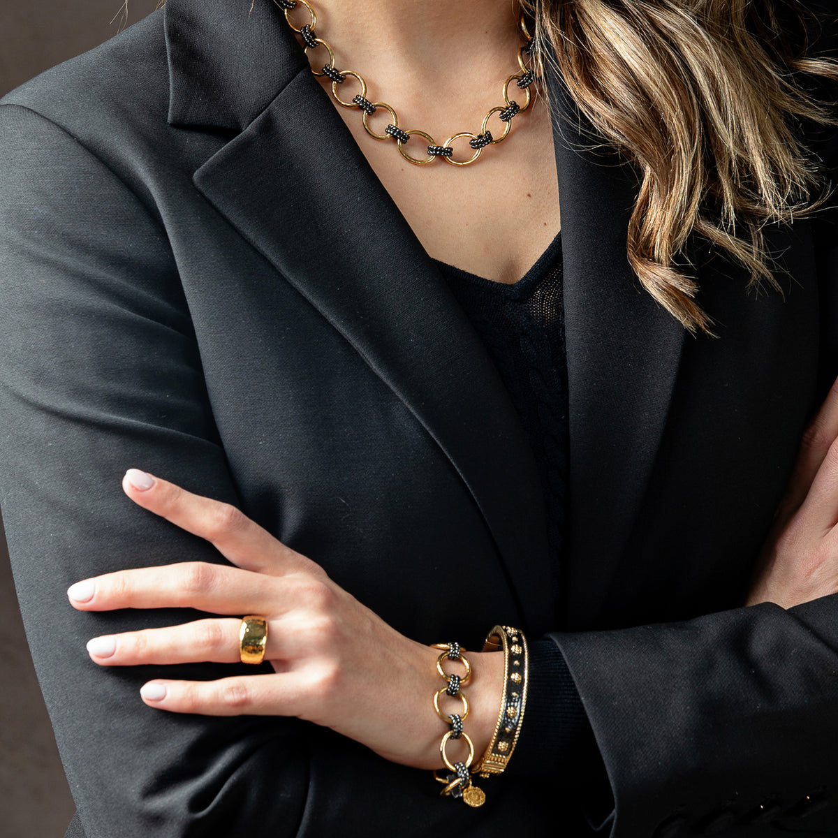 A close-up lifestyle shot of a woman wearing a gold and black beaded jewelry set, including a chunky link necklace, a ring, and a bracelet, styled with a black blazer.