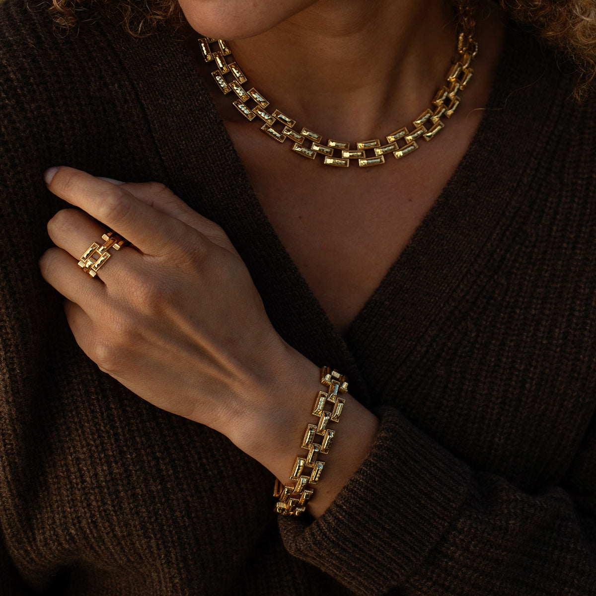 A close-up lifestyle shot of a model wearing a matching gold jewelry set, including a geometric link chain necklace, a matching bracelet, and a ring, styled with a dark brown knit cardigan.