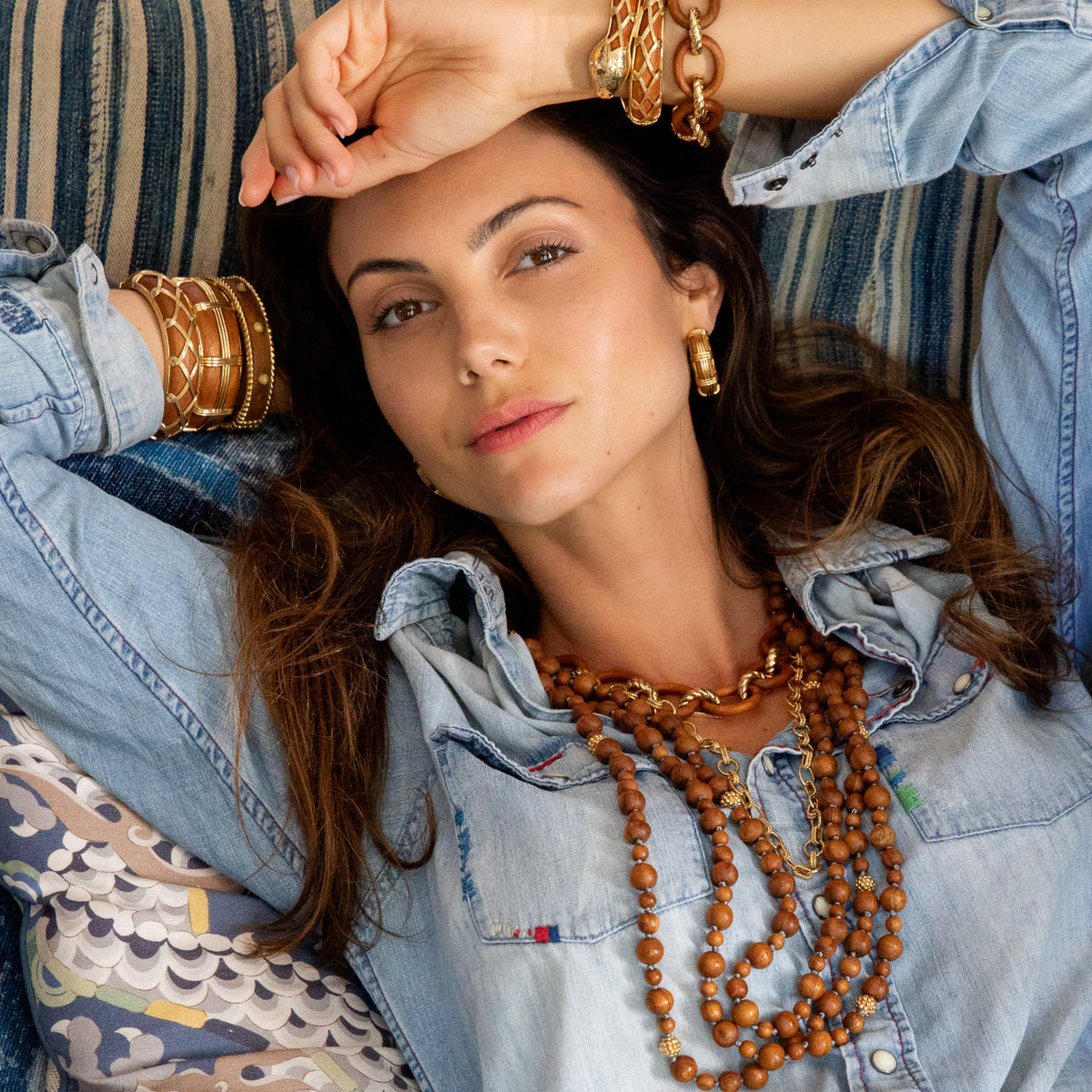 A woman wearing layered gold jewelry, including a chunky chain bracelet, bangles, and a multi-strand wooden bead necklace, lying on a striped couch.