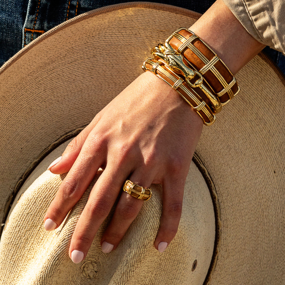 A close-up of a person's hand resting on a beige straw hat, showcasing a gold geometric ring and a stack of gold and brown geometric bangles on the wrist.