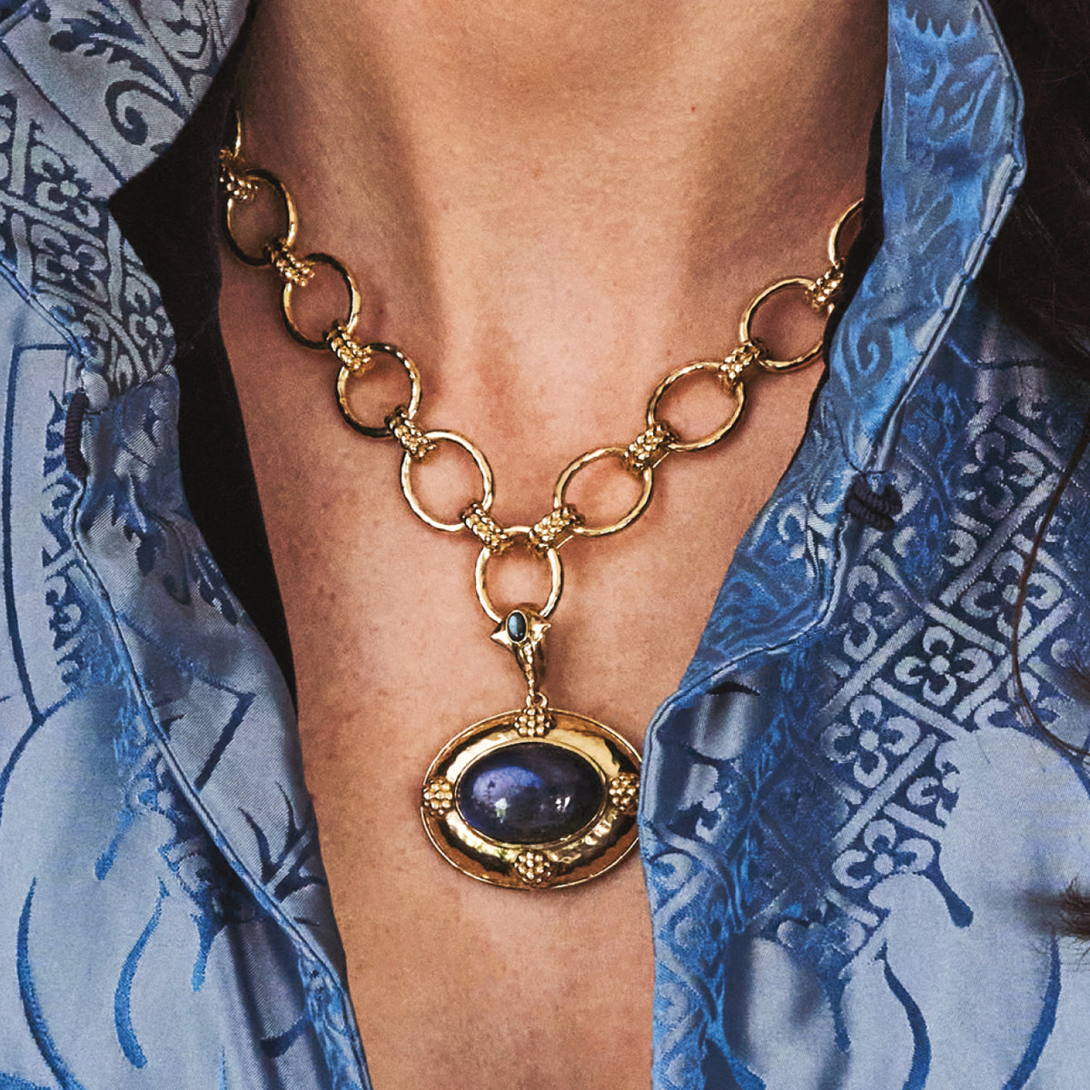 A close-up of a person wearing a gold-toned link necklace with a large oval pendant featuring a dark blue stone, styled with a blue patterned shirt.