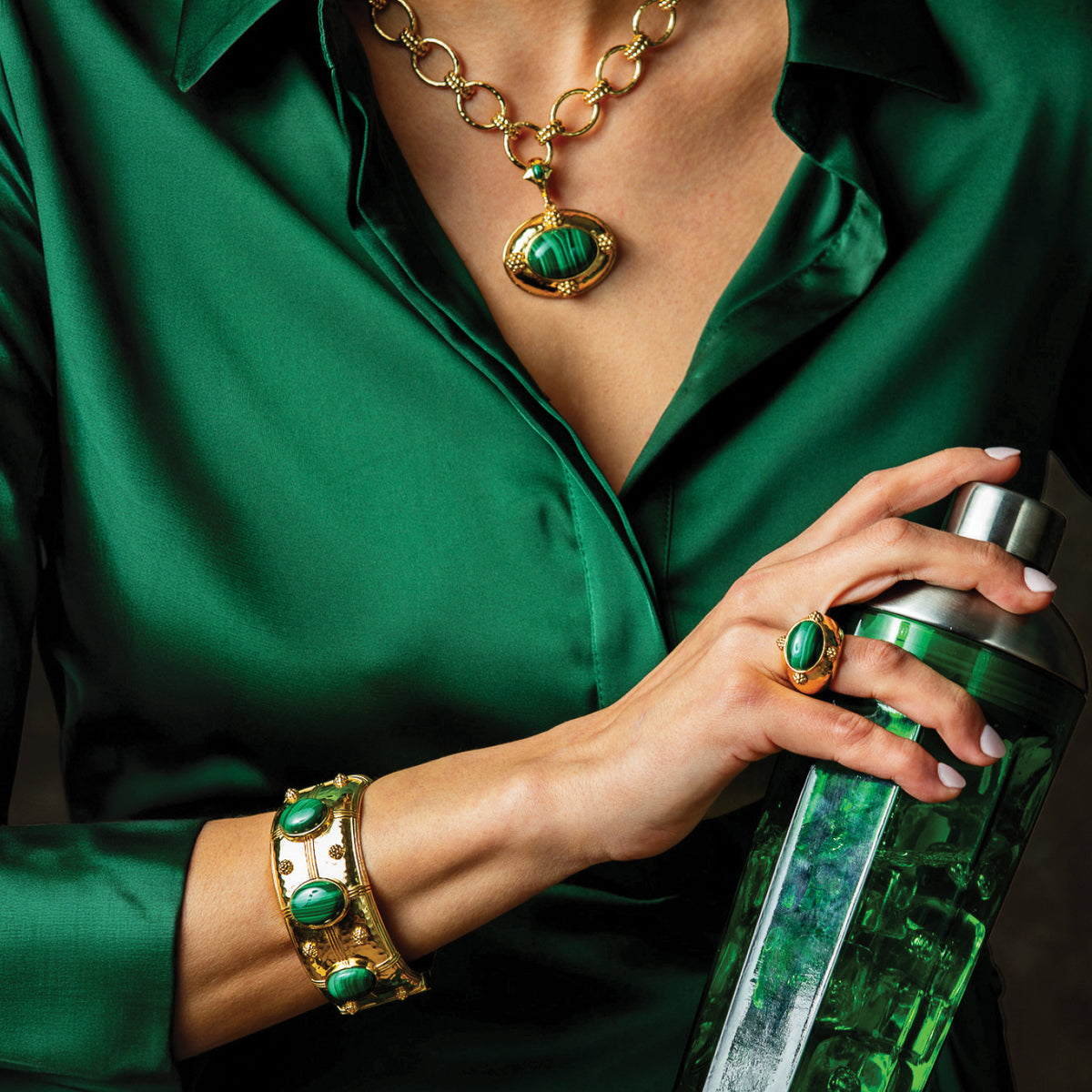 A close-up lifestyle shot of a person wearing a matching green silk blouse and gold jewelry with green stones, holding a green textured bottle. Visible jewelry includes a gold chain necklace with a green pendant, a gold ring, and a gold bracelet.