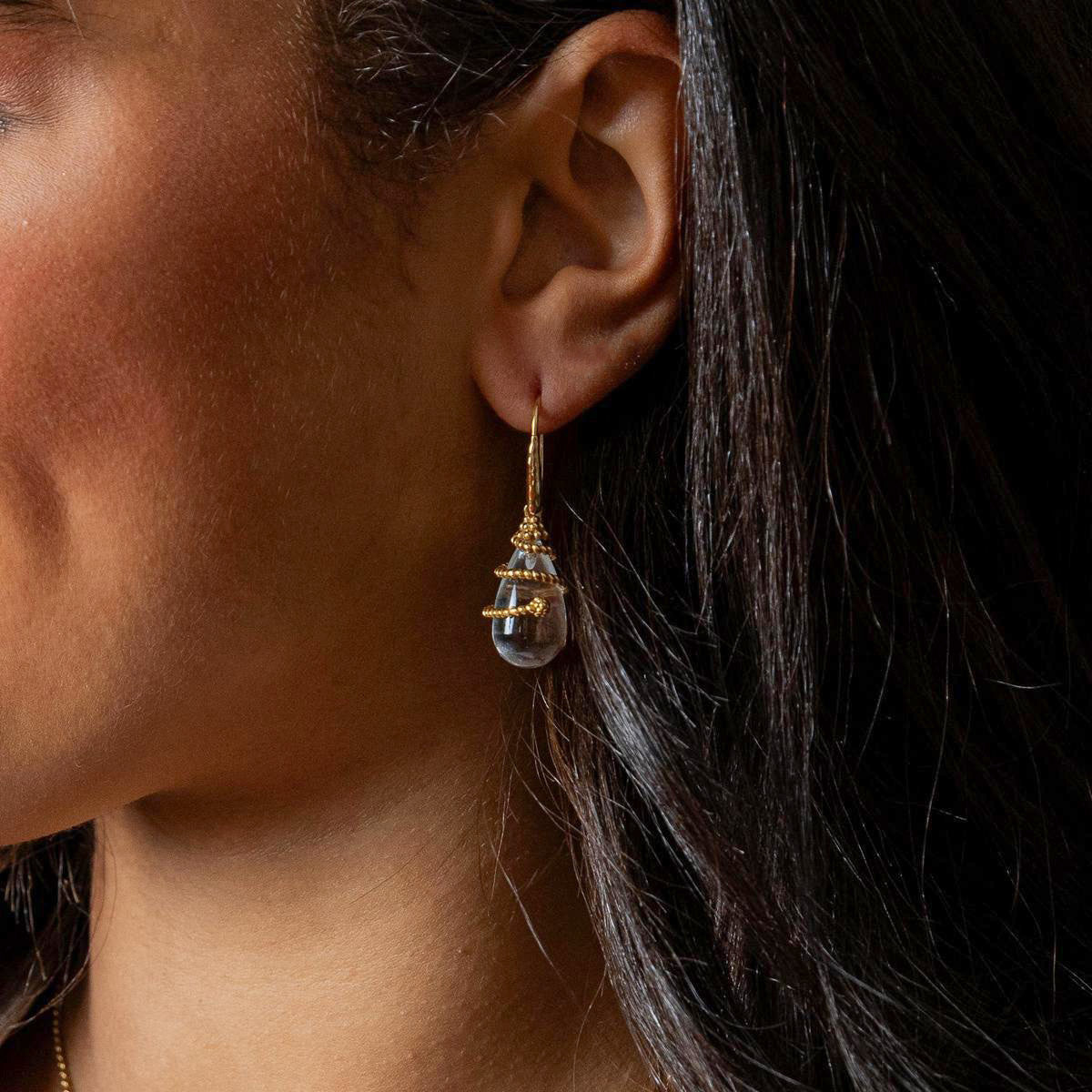 A close-up side profile of a woman wearing a delicate gold dangle earring with a clear teardrop-shaped gemstone.