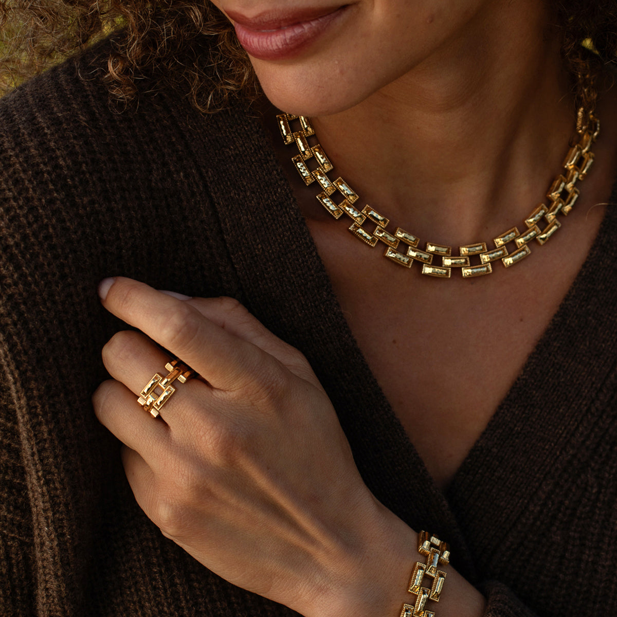 A close-up lifestyle shot of a model wearing a gold geometric link necklace, matching ring, and bracelet, paired with a dark brown knit cardigan.