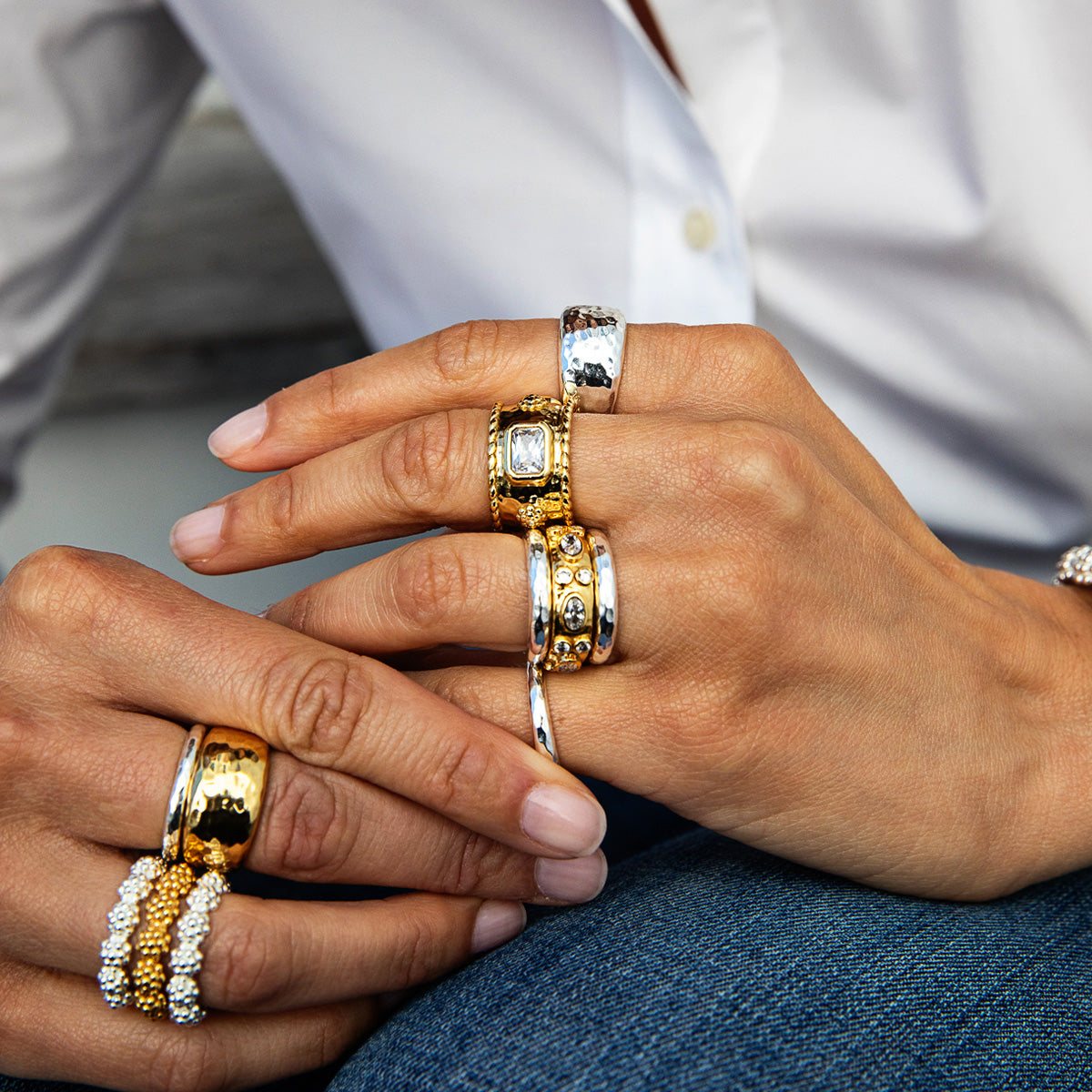 Close-up of a person's hands wearing multiple gold and silver rings, including one with a square-cut clear stone and others with textured and striped details, styled with a white shirt and blue jeans.
