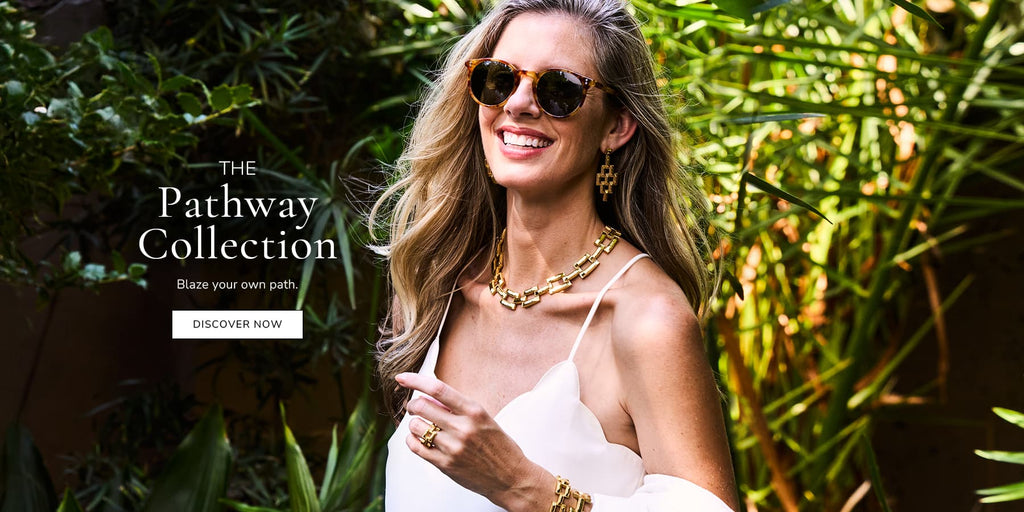 Woman in sunglasses and a white dress with gold jewelry against a green foliage background, promoting 'The Pathway Collection'.