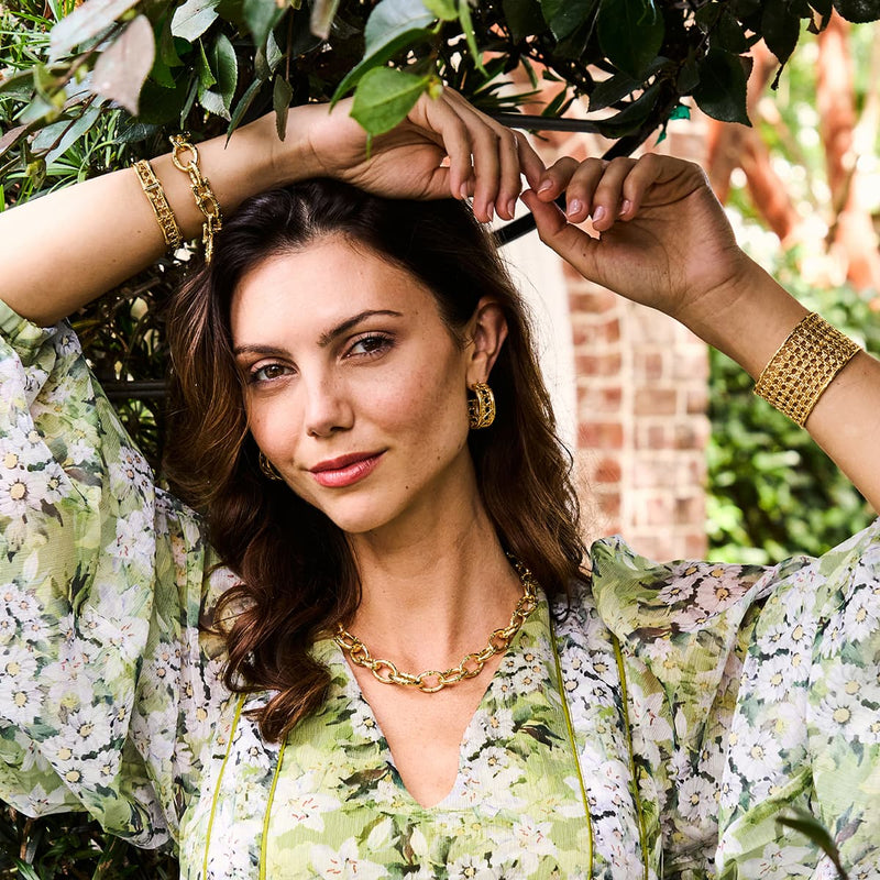 Woman in a green floral shirt with her arms slightly over her head wearing golden bracelets, necklace, and earrings
