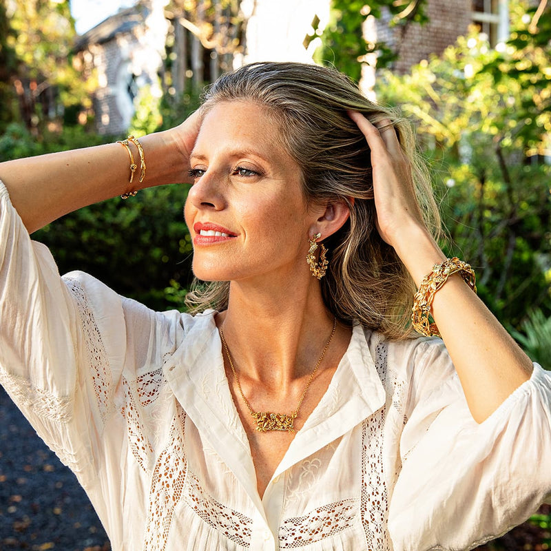 Woman in a white shirt running her hands through her hair, smiling and looking off into the distance. Wearing golden floral necklace, earrings, and bracelets