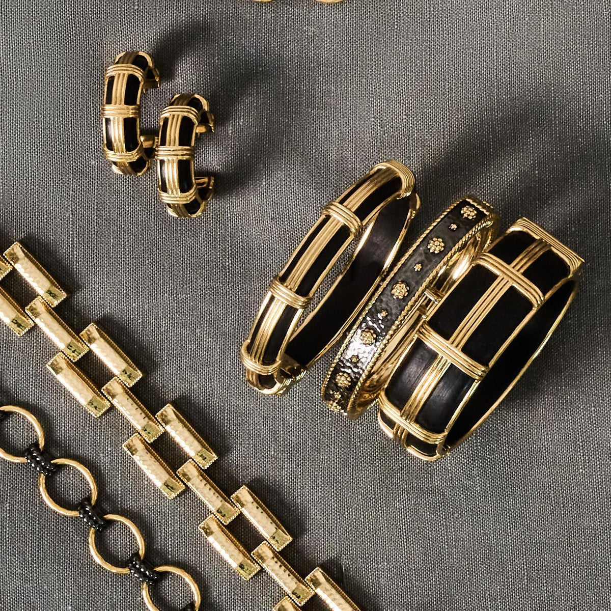 A jewelry set featuring gold-toned metal pieces with black enamel inlay, including two cuff bracelets, two hoop earrings, and a chain bracelet, arranged on a textured gray fabric background.