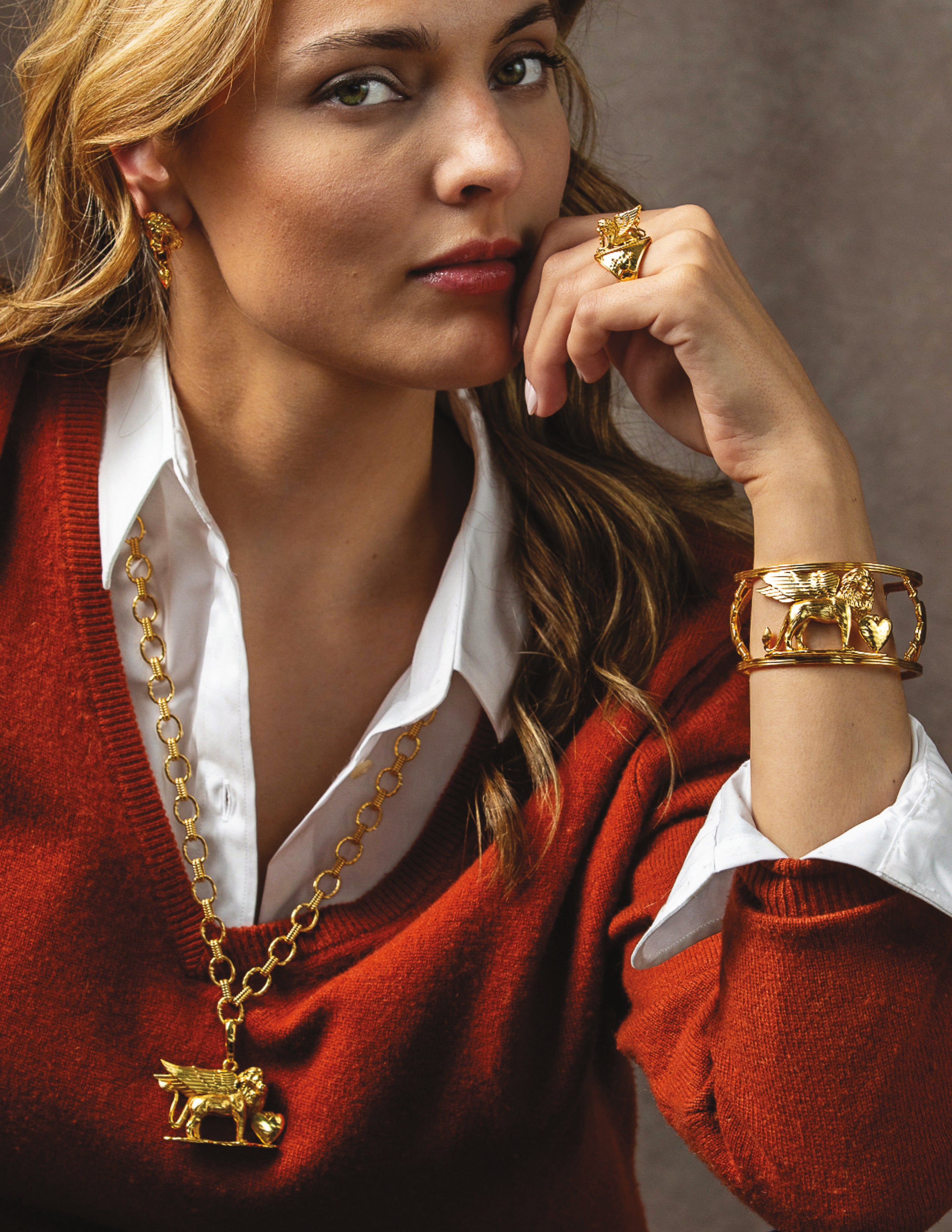 A close-up lifestyle shot of a woman wearing gold jewelry, including a moose pendant necklace, a ring, and a cuff bracelet, styled with a red sweater and white collared shirt against a neutral background.
