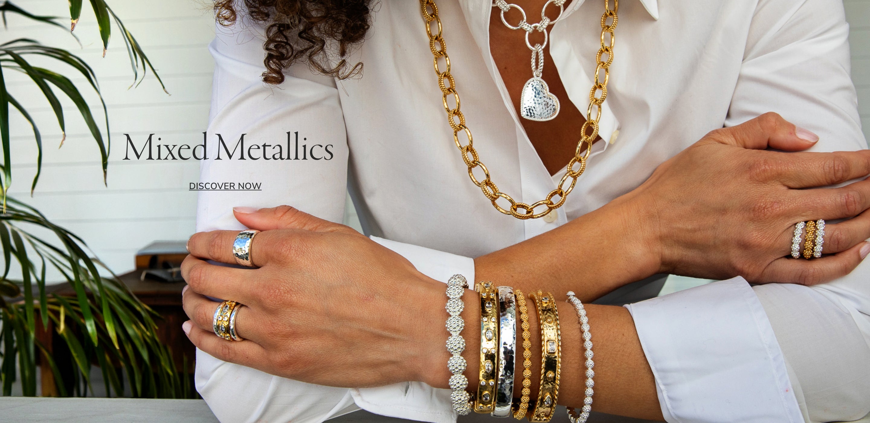 A person wearing mixed metallic jewelry, including layered gold and silver necklaces, rings, and bracelets, styled with a white shirt and a white paneled wall background. MIXED METALLICS. DISCOVER NOW.