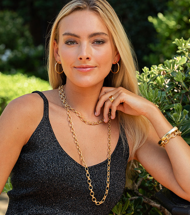 Woman wearing gold jewelry outdoors with greenery in the background