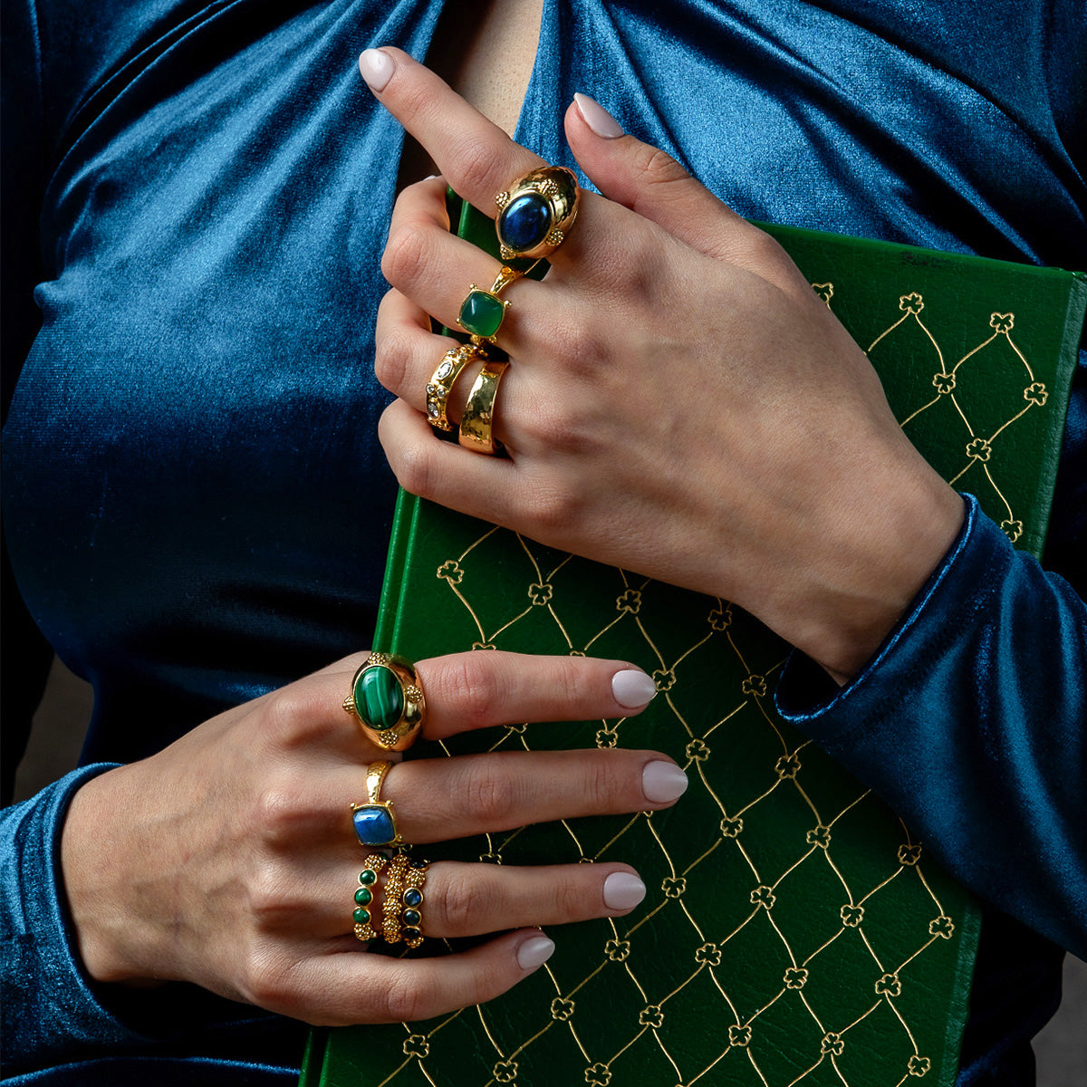 A close-up of a person wearing multiple gold rings with green and blue stones, holding a green book with a gold lattice pattern against a dark background.