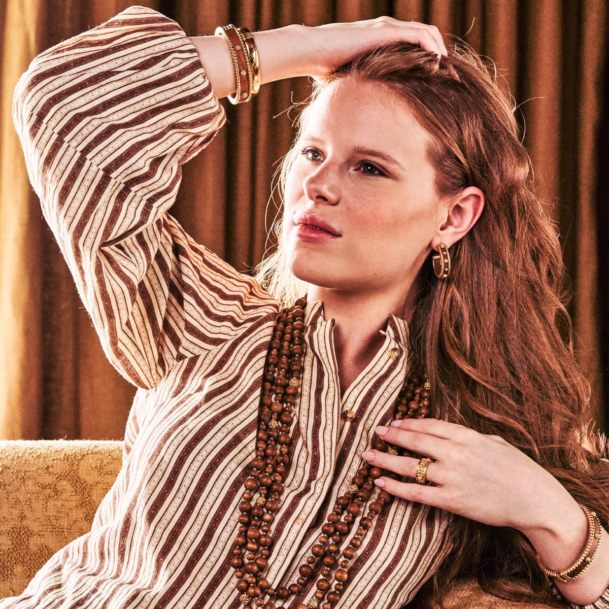 Woman sitting on a couch wearing a striped dress and multiple necklaces, with a warm-toned background.