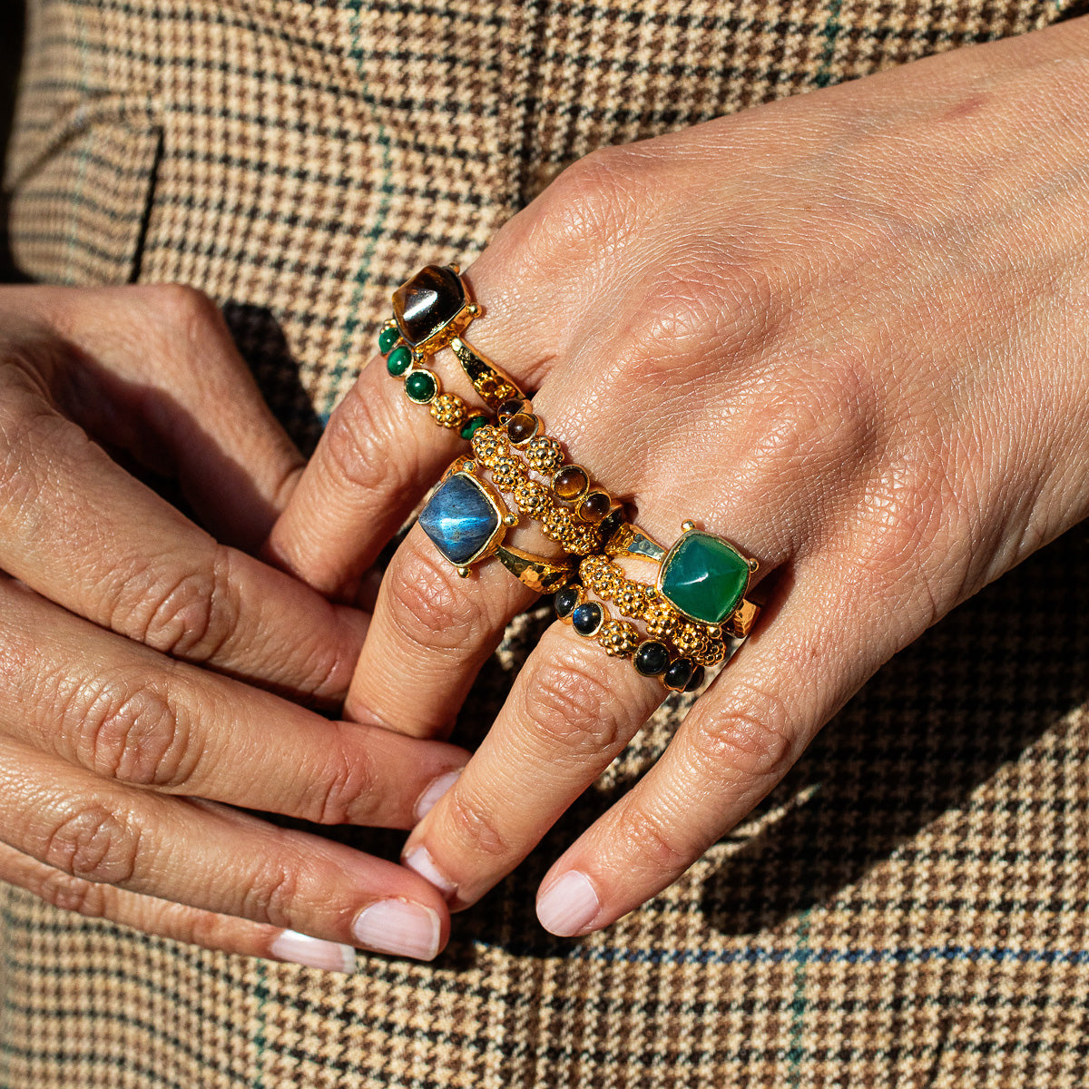 A close-up of a person's hands wearing multiple ornate gold rings with green and blue stones, set against a brown and black houndstooth patterned jacket.