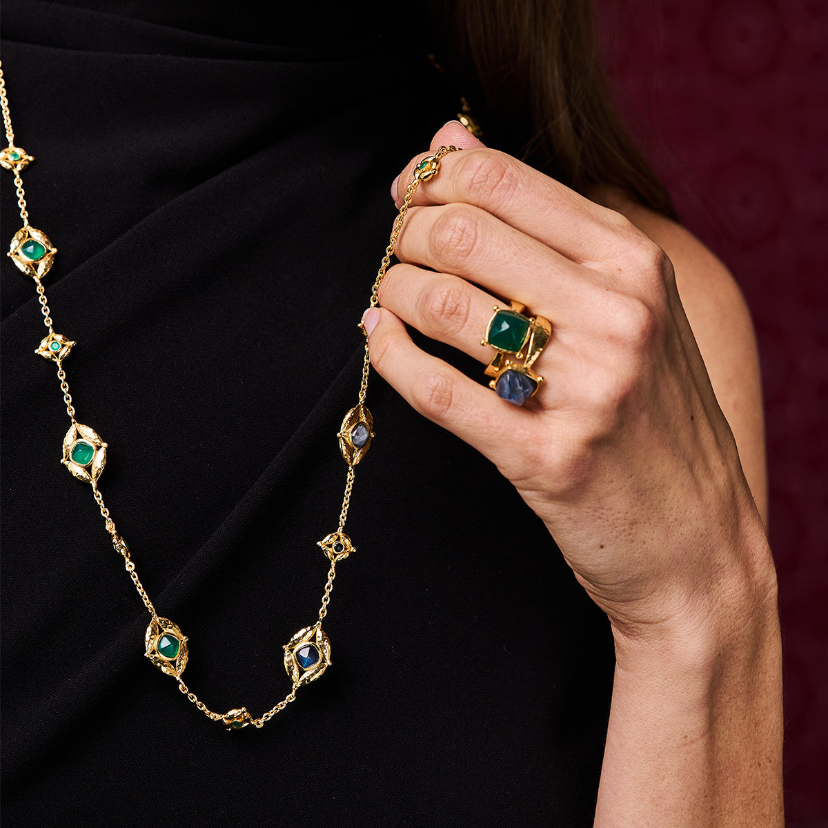 Gold necklace with green and blue gemstones held by a hand against a dark background