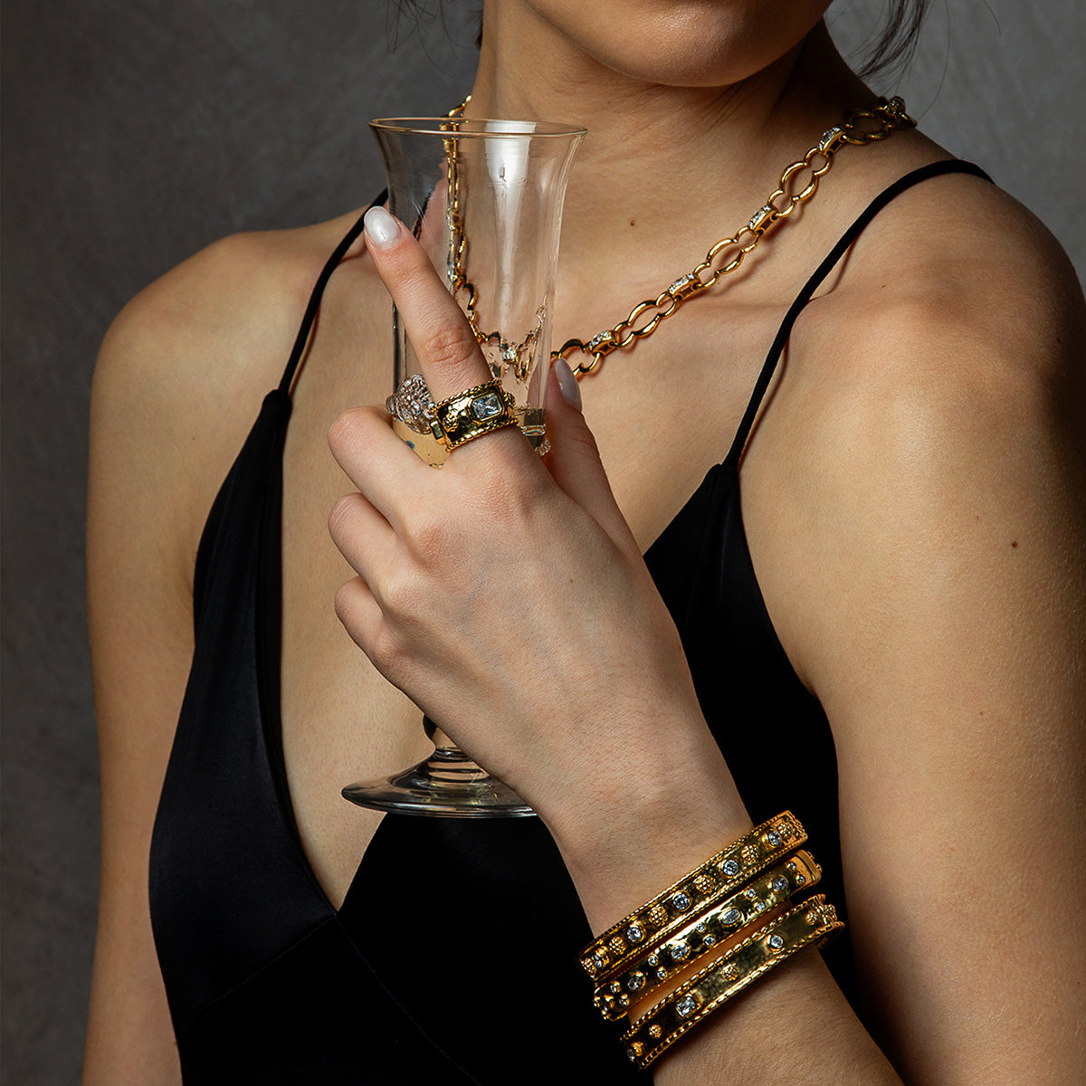 A close-up lifestyle shot of a model wearing a gold chain necklace, a statement ring, and stacked gold bangles with clear crystals, holding a champagne flute while wearing a black dress against a dark gray background.